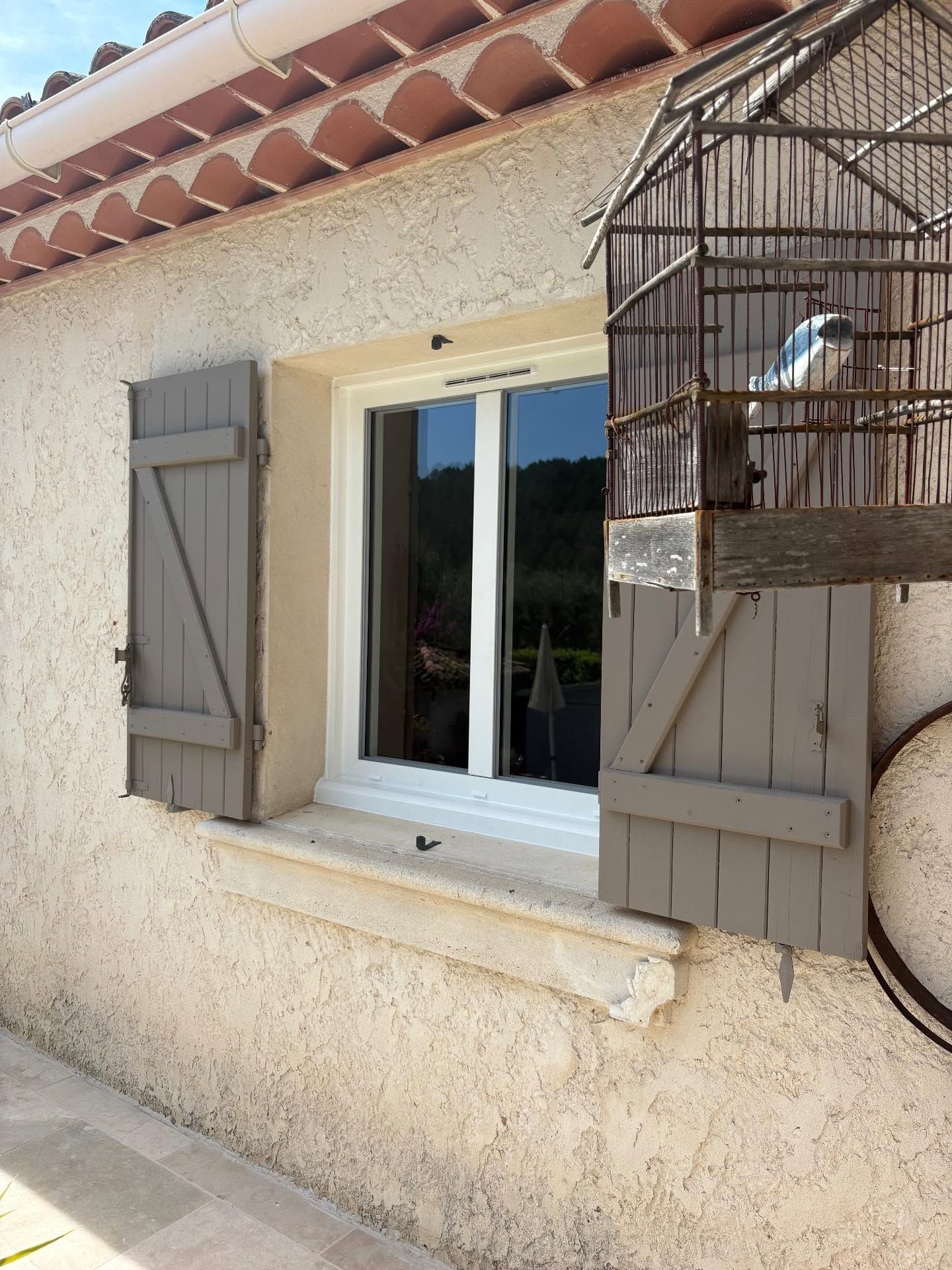 Extérieur d'une maison blanche avec une fenêtre, des volets gris et une cage à oiseaux sur le côté droit.