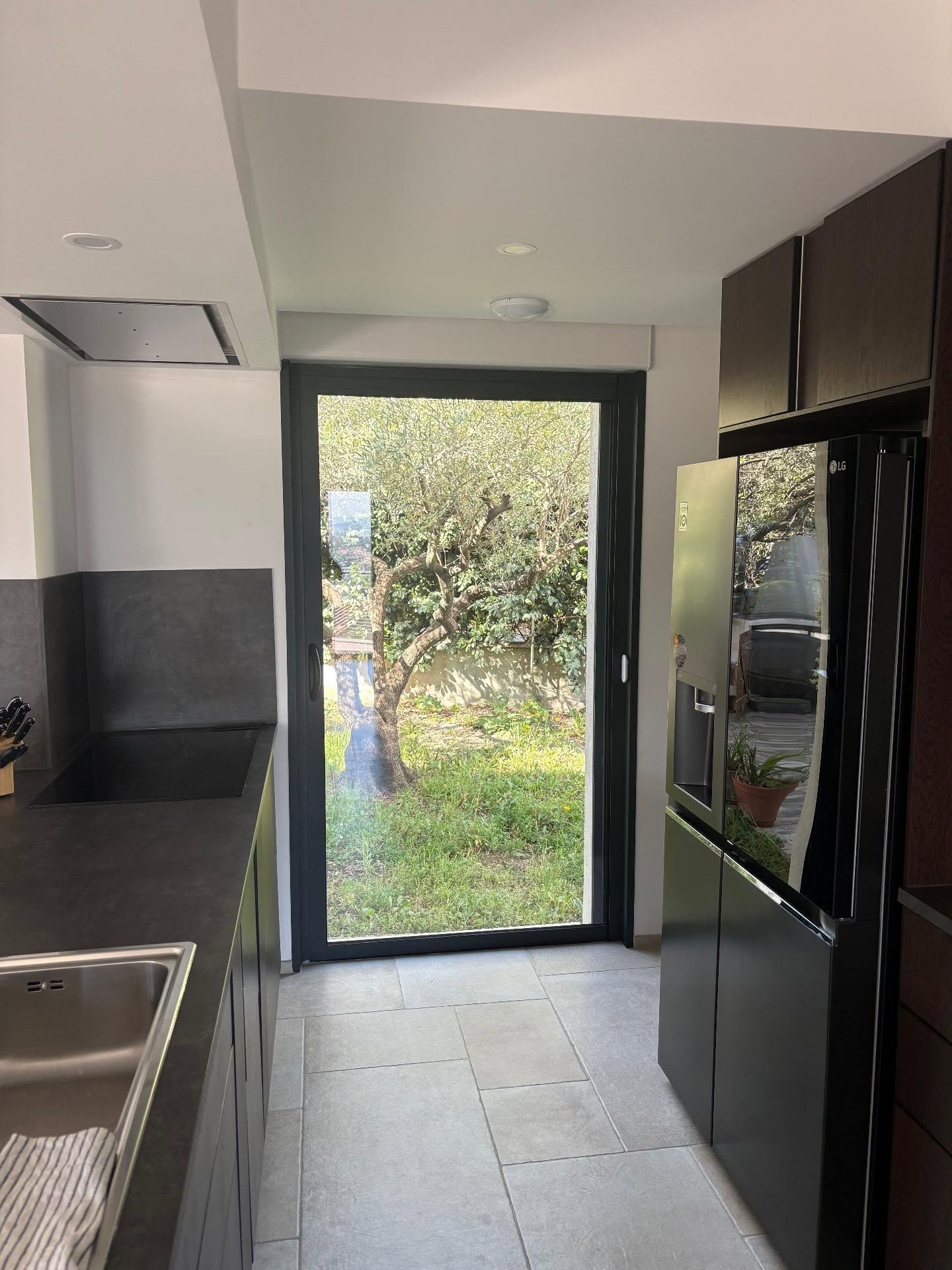 Cuisine moderne étroite avec placards noirs, sol carrelé blanc et porte vitrée donnant sur un jardin verdoyant.