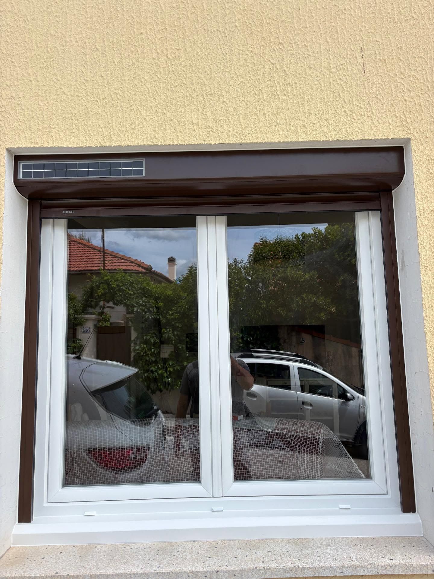 Fenêtre battante à cadre blanc dans un mur extérieur beige, reflétant une rue et une voiture garée.