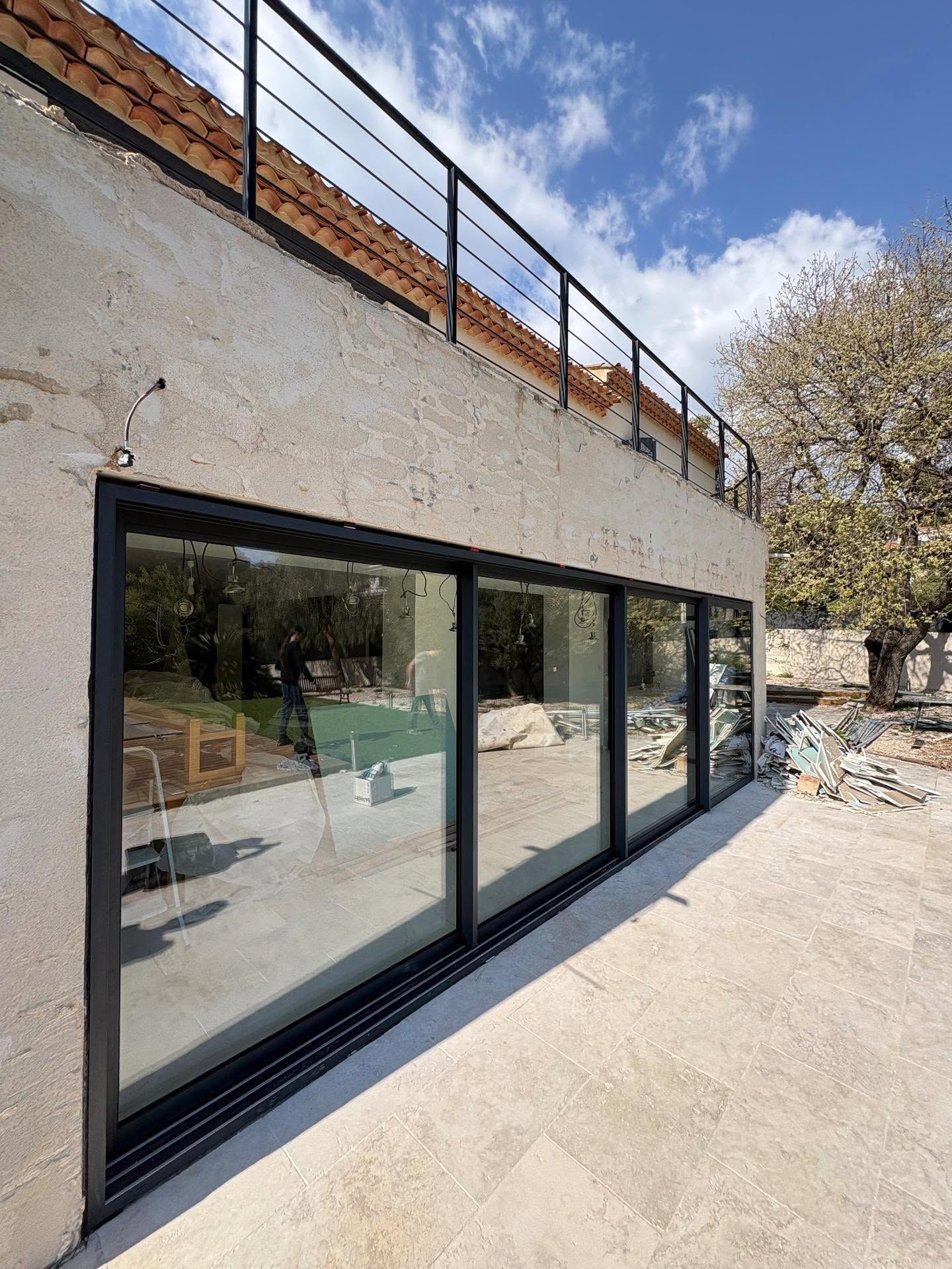 Extérieur d'une maison moderne avec de grandes portes vitrées à cadre noir et une balustrade sur le toit, sous un ciel bleu.
