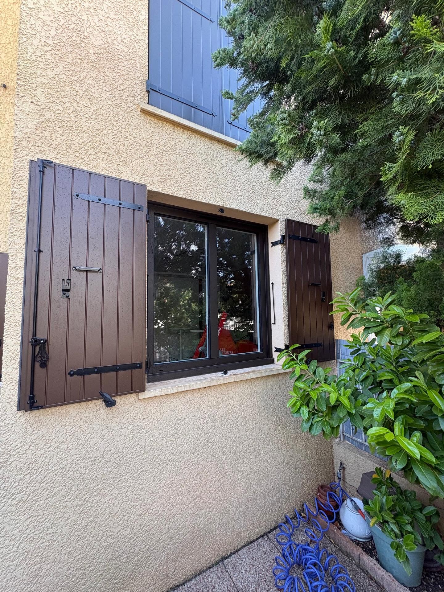 Fenêtre de maison avec volets marron foncé ouverts, à côté de verdure et de plantes en pot