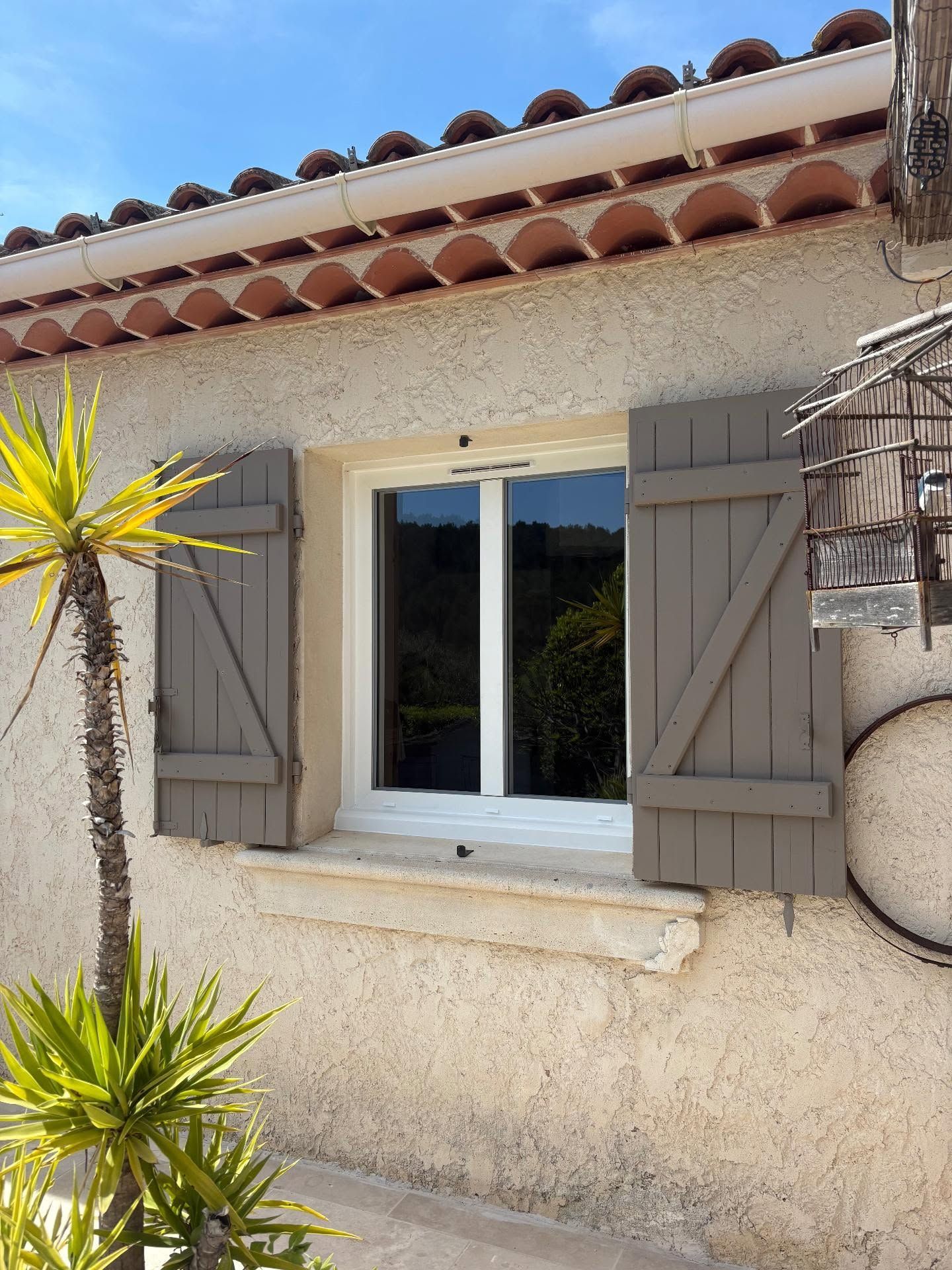 Fenêtre d'une maison en stuc avec volets beiges, cadre blanc et toit de tuiles en terre cuite.
