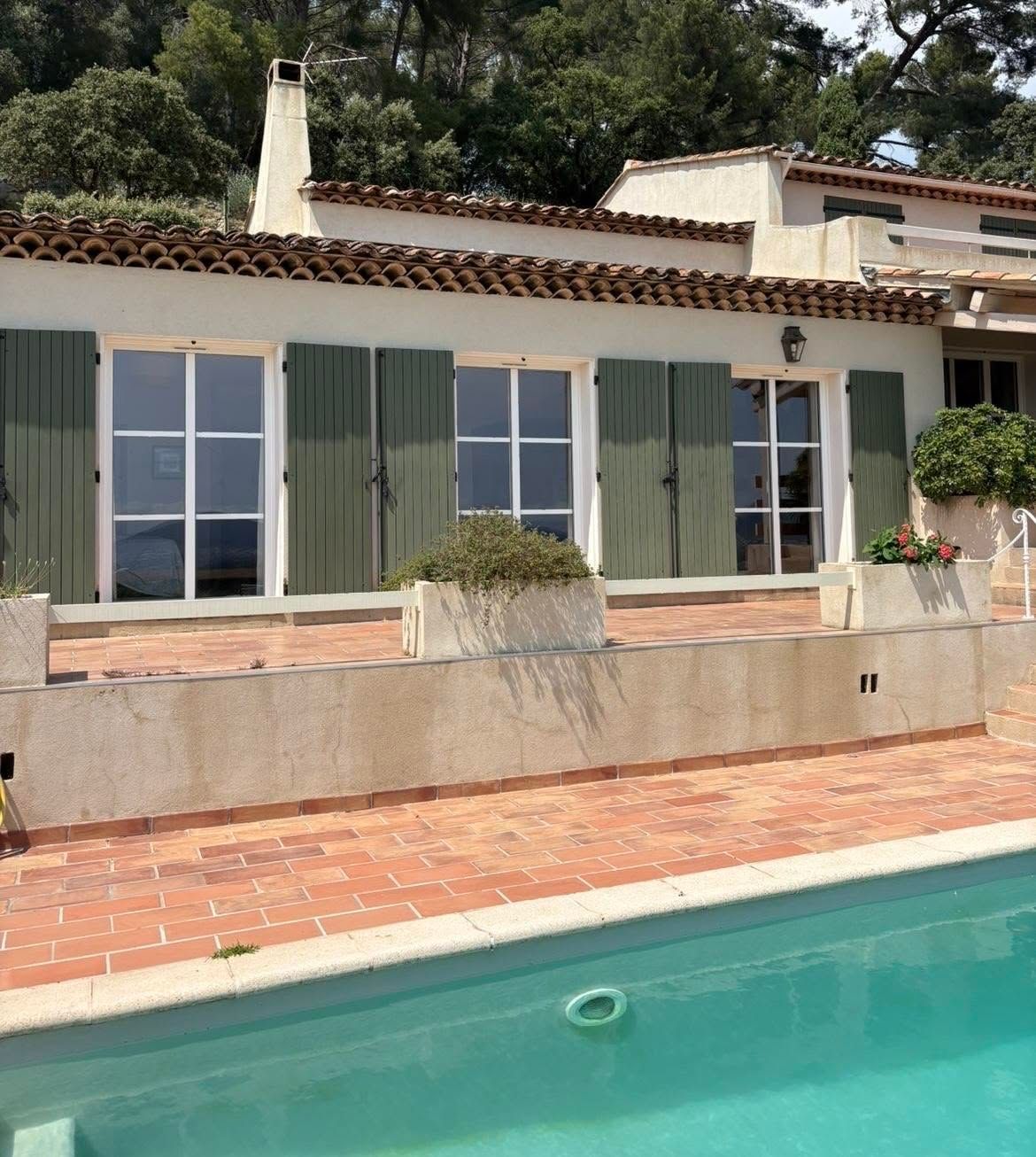 Maison blanche aux volets verts, à côté d'une piscine et d'une terrasse en briques.