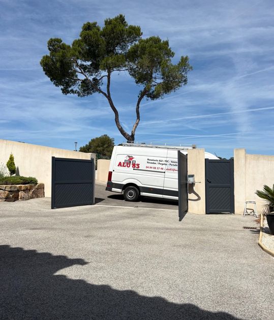 Une camionnette de service blanche garée dans une allée privée fermée par un portail, sous un grand arbre, par une journée ensoleillée.