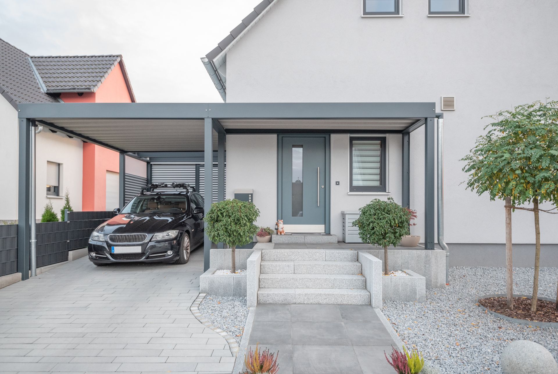 Extérieur d'une maison moderne avec abri voiture, voiture noire garée, marches d'entrée et aménagement paysager en gravier.