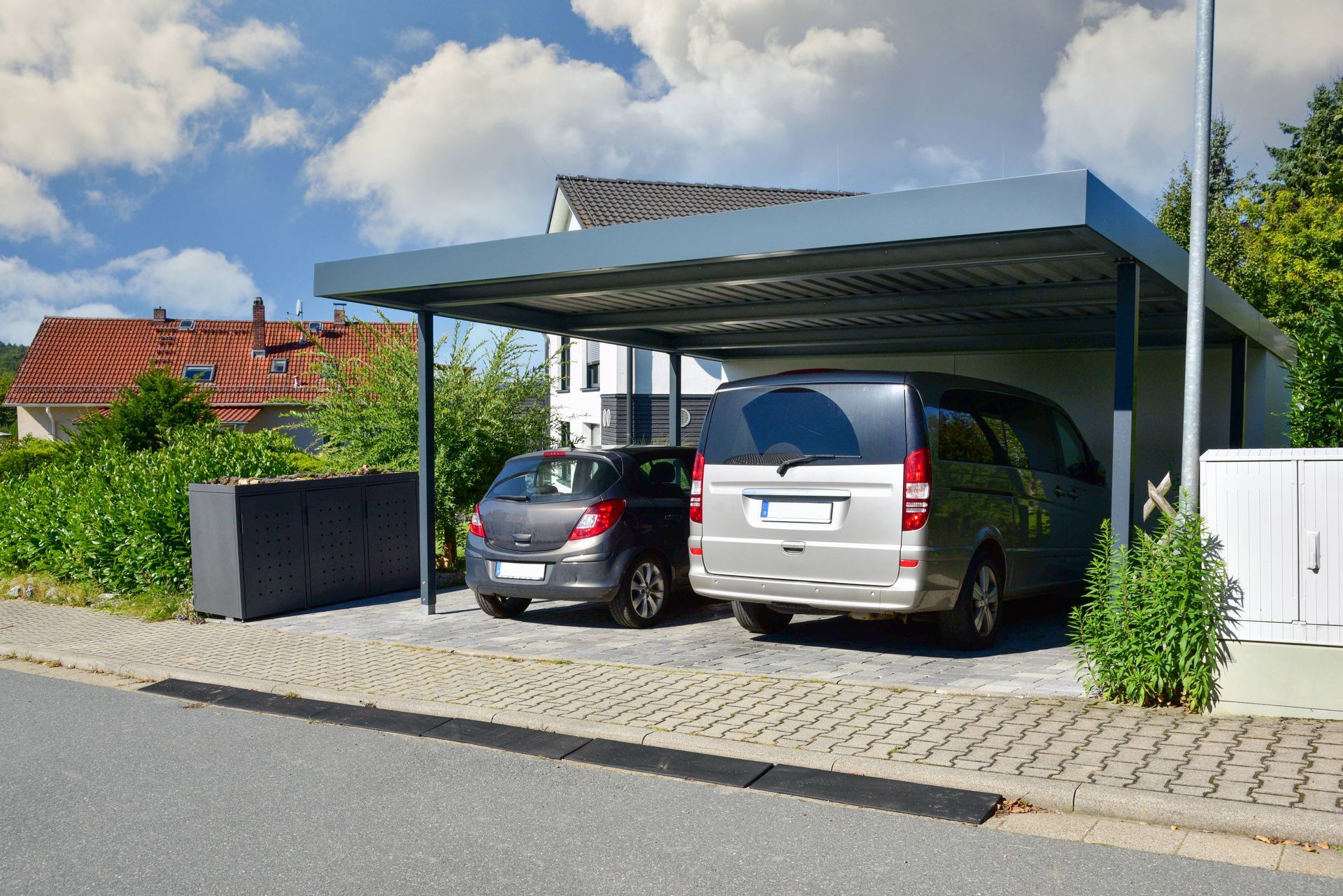Deux voitures garées sous un abri métallique au bord d'une rue d'un quartier de banlieue
