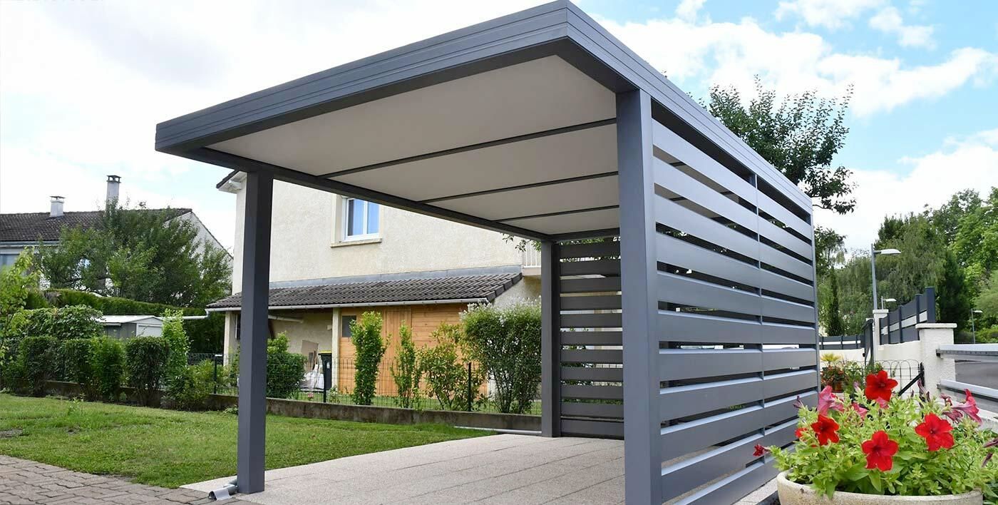 Allée couverte à côté d'une maison avec un abri voiture en béton, une clôture et des arbres de jardin.