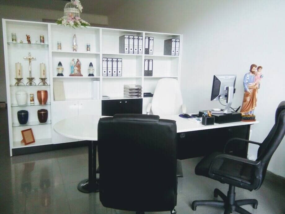 Escritório com mesa, prateleiras com objetos religiosos e duas cadeiras de escritório pretas.