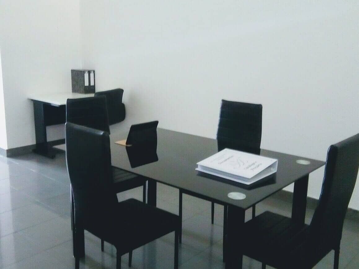 Um pequeno escritório com uma mesa de vidro preta e quatro cadeiras pretas. 