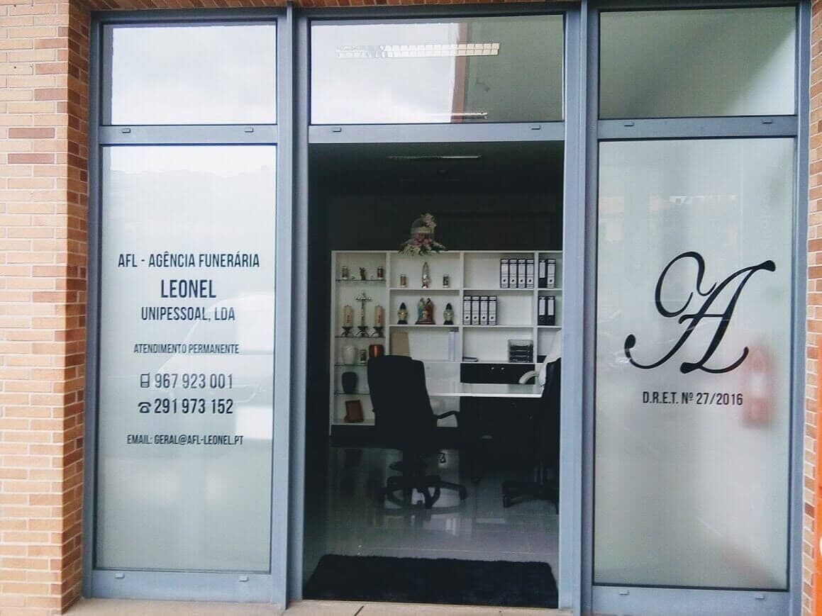 Fachada de um prédio comercial com janelas de vidro fosco exibindo nomes de empresas e informações de contato. Porta aberta.