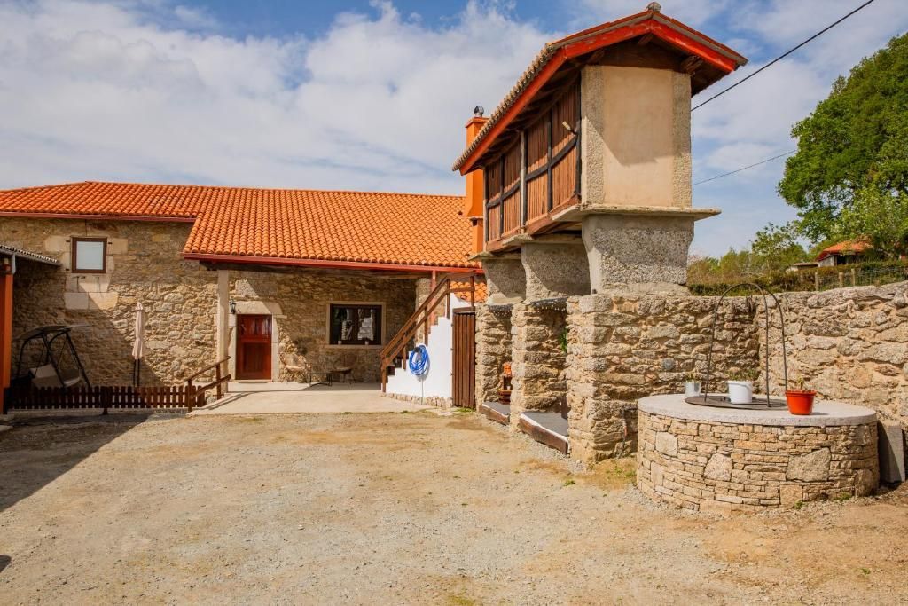 Patio de una casa de campo de piedra con techo de tejas rojas, torre blanca y pozo de piedra bajo un cielo azul.