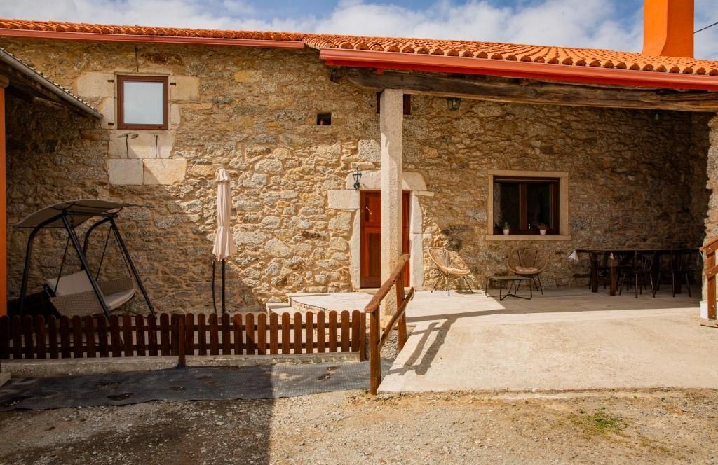Patio de una casa de campo de piedra con techo de tejas rojas, valla de madera y porche sombreado bajo un cielo soleado.