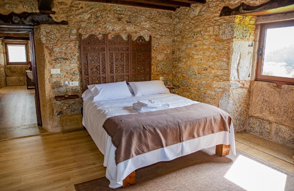 Habitación rústica de piedra con cama de madera, sábanas blancas y luz natural que entra por dos ventanas.