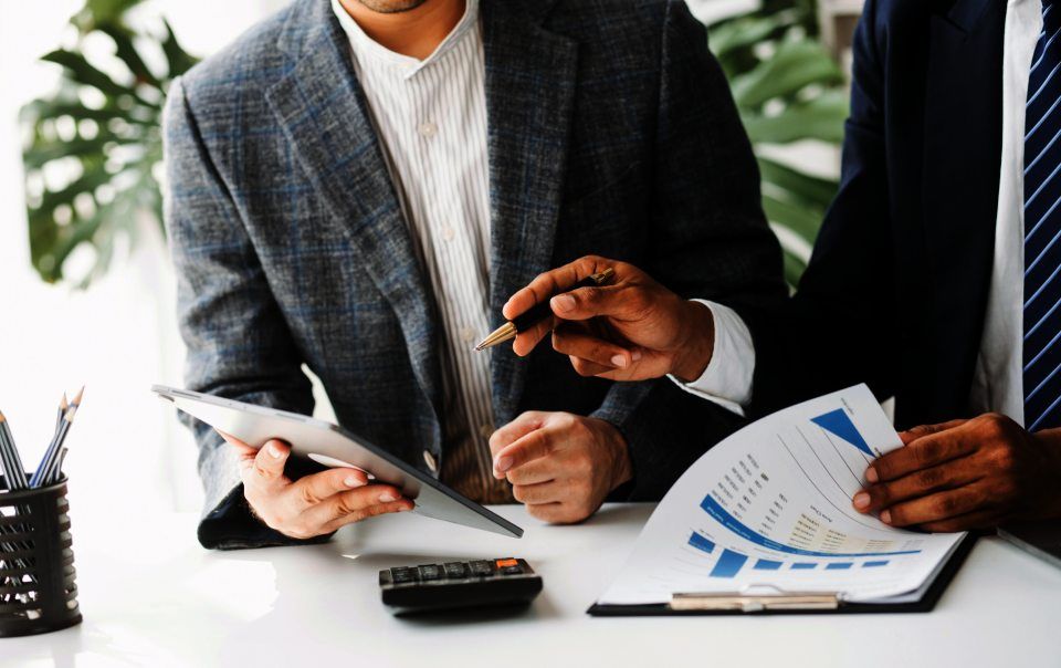 Zwei Männer sitzen an einem Tisch und schauen auf Papiere und ein Tablet.