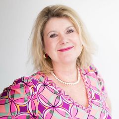 Lächelnde blonde Frau, trägt ein gemustertes rosa/lila Oberteil und eine Perlenkette; weißer Hintergrund.