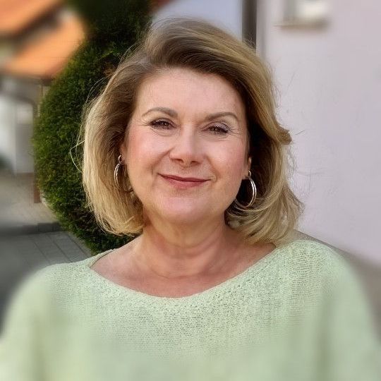 Frau mit blondem Haar lächelt im Freien neben einer Steinmauer; sie trägt ein buntes Oberteil mit Blumenmuster.