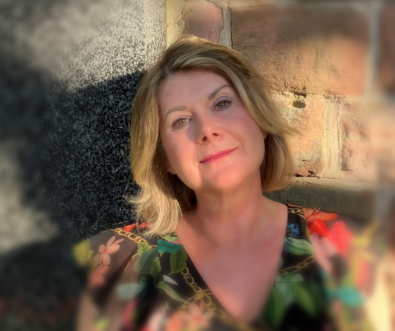 TrostWorte Patricia Rind Was macht eine freie Trauerrednerin? Aufgaben, Ablauf und Haltung bei einer Trauerfeier in der Metropolregion Rhein Neckar verständlich erklärt. Frau mit blonden Haaren, lächelnd, im Freien vor einer Steinmauer, trägt ein geblümtes Oberteil.