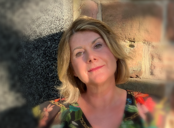 Frau mit blondem Haar lächelt im Freien neben einer Steinmauer; sie trägt ein buntes Oberteil mit Blumenmuster.