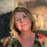 Frau mit blonden Haaren lächelt im Freien. Trägt geblümtes Oberteil, lehnt an einer Steinmauer, Sonnenlicht.