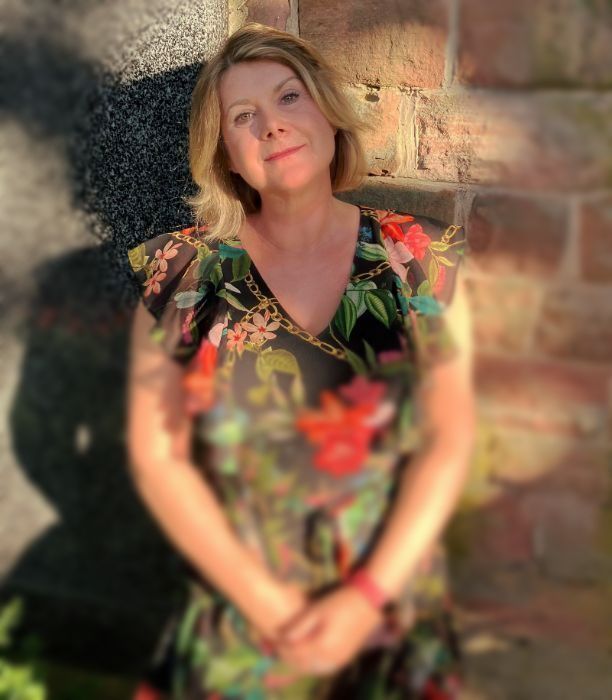 Eine Frau mit blondem Haar lächelt, trägt ein geblümtes Kleid und steht im Sonnenlicht vor einem Backsteingebäude.