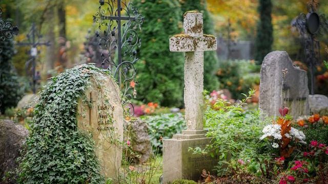 Friedhof mit moosbedeckten Grabsteinen und einem zentralen Kreuz, umgeben von Laub und Blumen.
