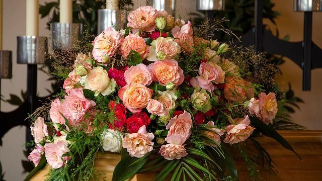 Arrangement aus rosa und pfirsichfarbenen Rosen, Grünpflanzen und anderen Blumen auf einer Holzoberfläche.