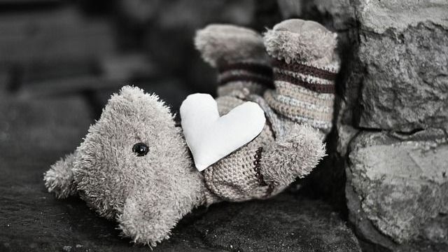 Teddybär mit einem weißen Herz auf der Brust, vor einer Steinmauer; einfarbig.