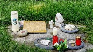 Grabstätte mit Gedenksteinen, darunter Kerzen, ein herzförmiger Stein und eine Gedenktafel, auf einer Rasenfläche.