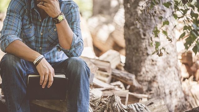 Mann in blau kariertem Hemd und Jeans sitzt im Freien, Kopf in der Hand, Buch in der Hand.