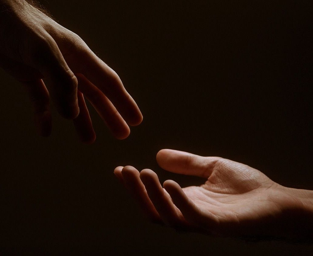 Zwei Hände, die sich vor einem dunklen Hintergrund nacheinander ausstrecken; die Finger berühren sich fast.