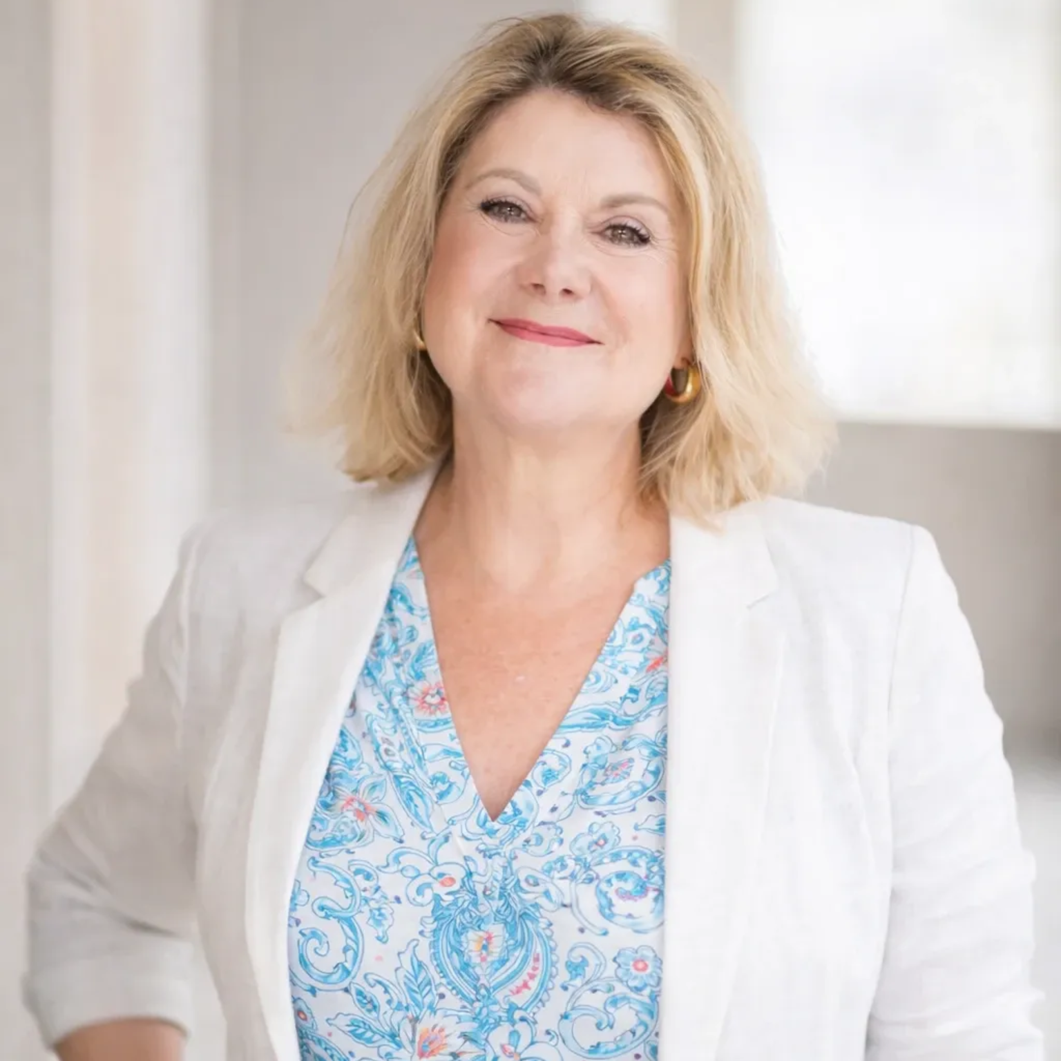 Professionelles Porträtfoto einer blonden Frau, die einen weißen Blazer über einem blau-weiß gemusterten Oberteil trägt.