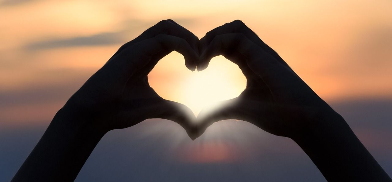 Hände, die ein Herz formen, zeichnen sich als Silhouette vor einem goldenen Sonnenuntergang ab.
