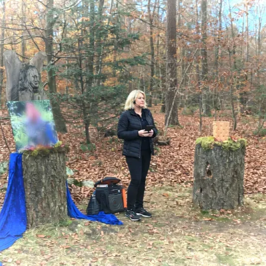 Eine Frau steht in einem Ruheforst zwischen zwei Baumstümpfen, einer blauem Stoff und einem Bild, auf dem anderen eine Urne