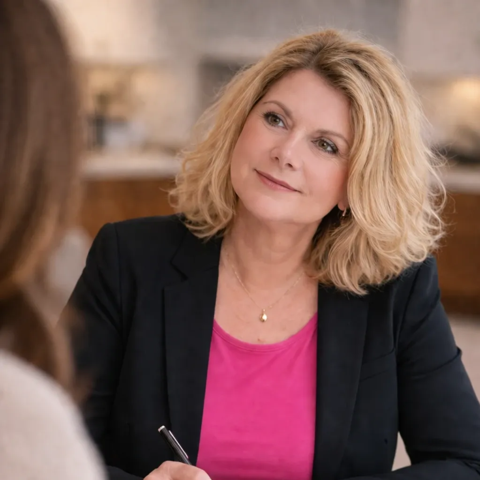 Frau in schwarzem Blazer und rosa Top sitzt in Besprechung am Tisch und hört aufmerksam zu; sie hält einen Stift in der Hand