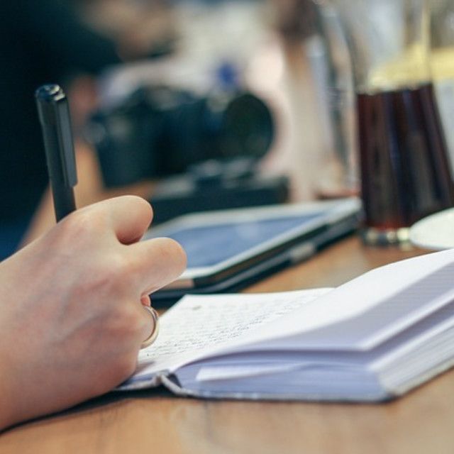 Jemand schreibt in ein Notizbuch, daneben stehen Getränke und ein Tablet auf einem Tisch.