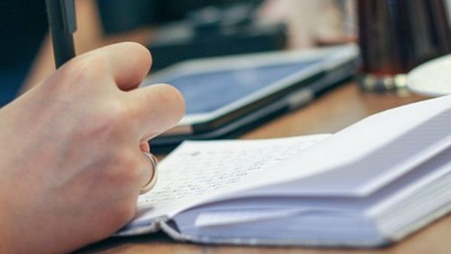 Person, die mit einem Stift in ein Notizbuch schreibt, im Hintergrund ein Tablet-Computer.