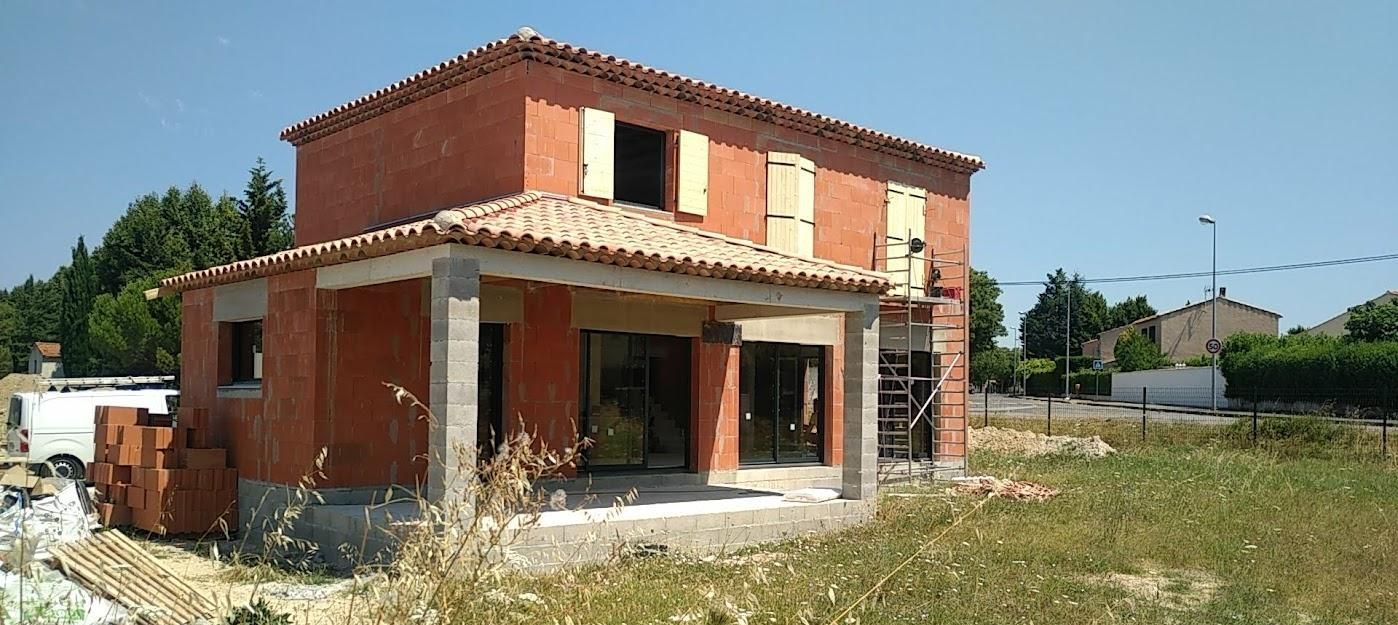 Maison en briques rouges partiellement construite, avec toit de tuiles et porche en béton inachevé.