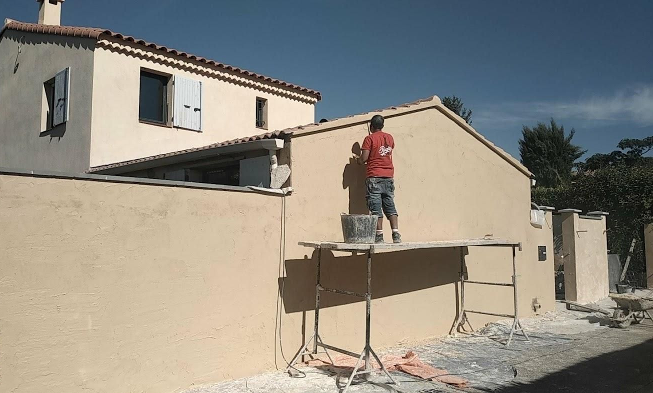 Un ouvrier en chemise rouge, debout sur un échafaudage, peint un mur en stuc beige à côté d'une maison.