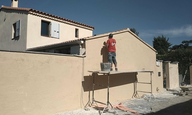 Un ouvrier en chemise rouge, debout sur un échafaudage, peint un mur en stuc beige à côté d'une maison.