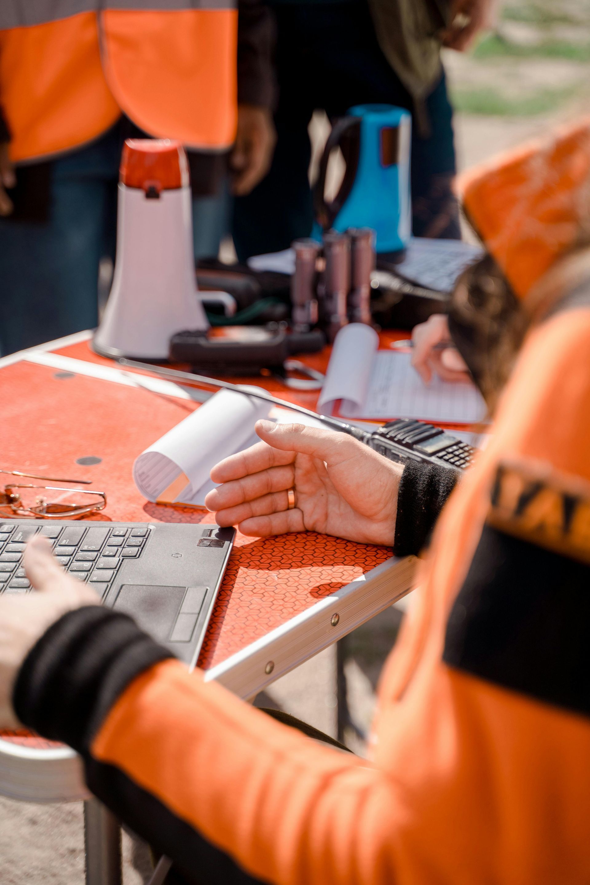 Des personnes portant des gilets de sécurité orange manipulent des outils et des documents à une table de travail extérieure