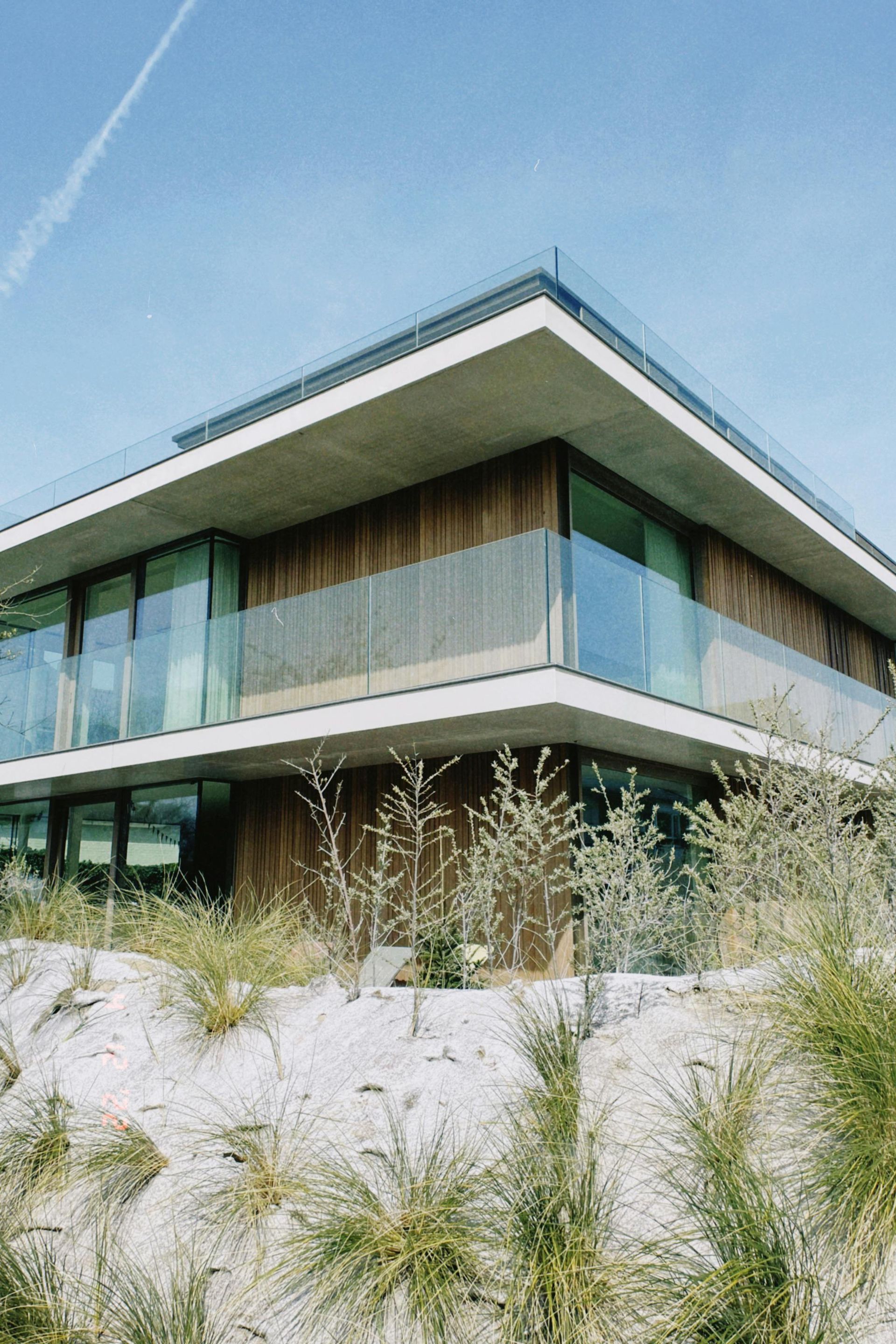 Maison moderne à deux étages avec de grandes baies vitrées et un toit plat, partiellement dissimulée par de hautes herbes.