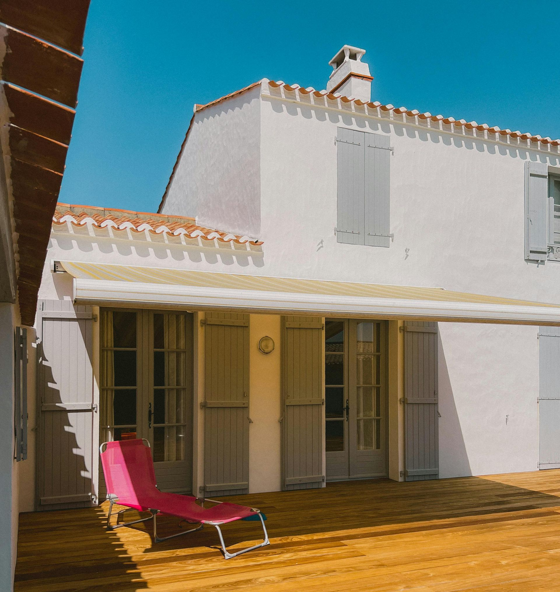 Patio ensoleillé avec façade de maison blanche, terrasse en bois, chaise longue rose et ciel bleu.