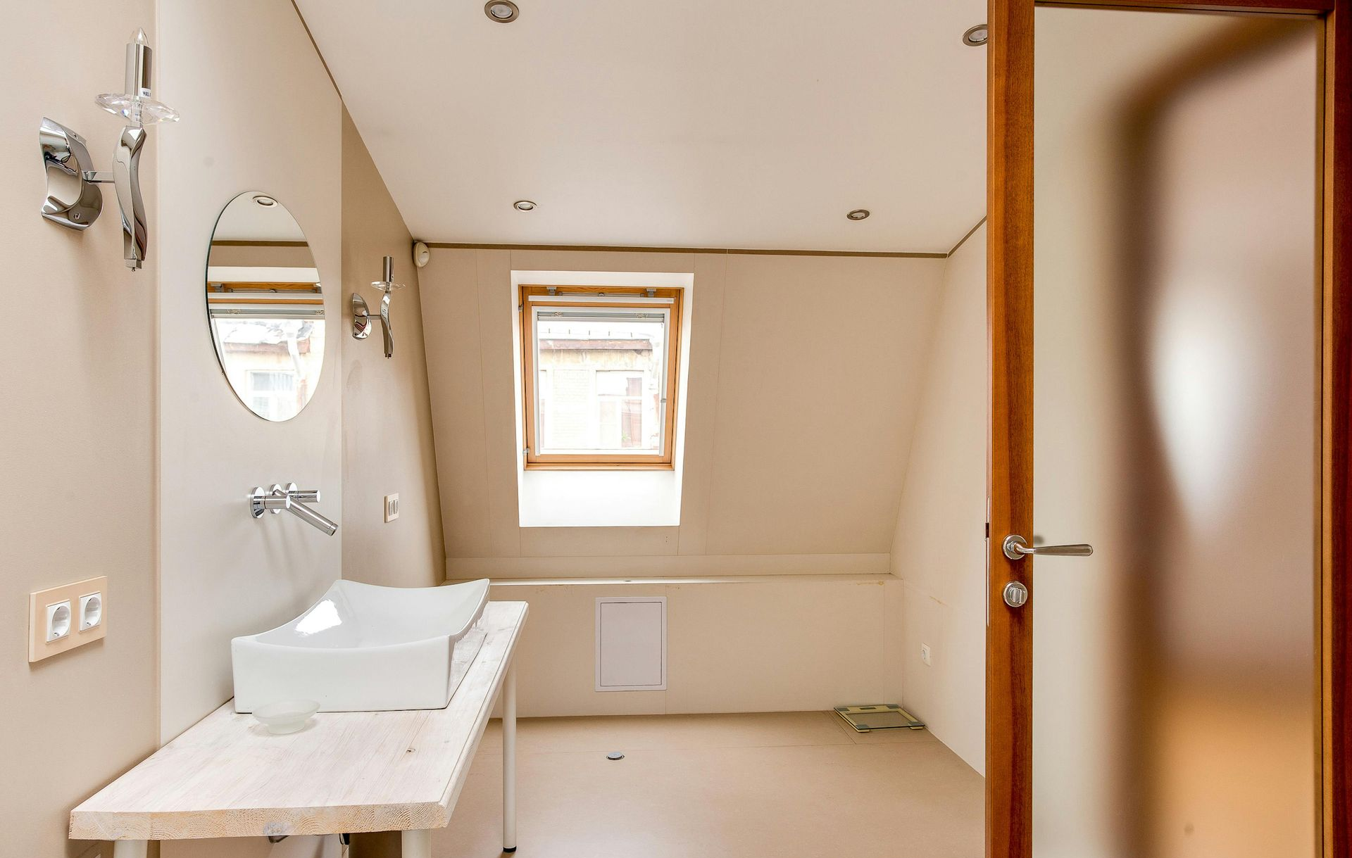 Salle de bains lumineuse avec lavabo blanc, miroir ovale, petite fenêtre et porte en bois ouverte.