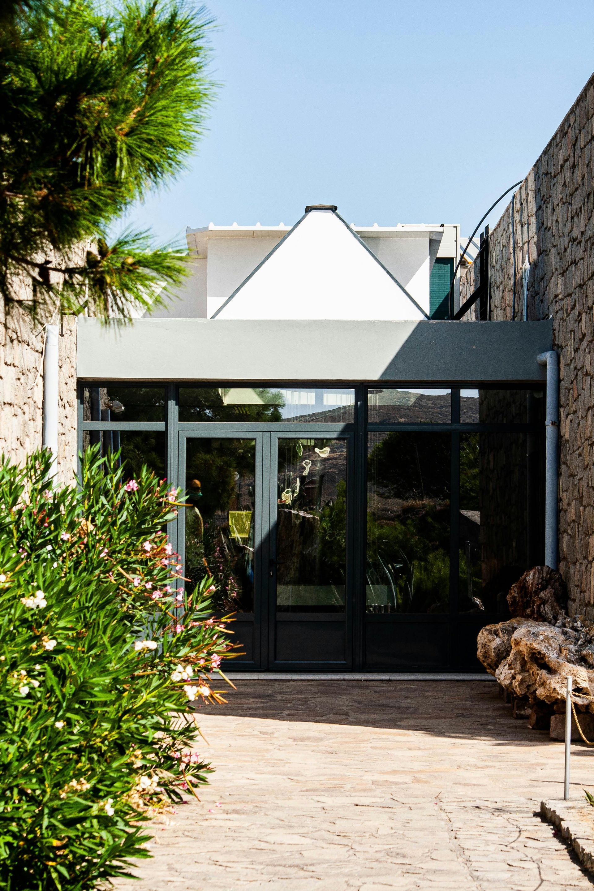 Entrée d'une maison moderne avec façade vitrée, toit triangulaire blanc, mur en pierre et allée paysagée.