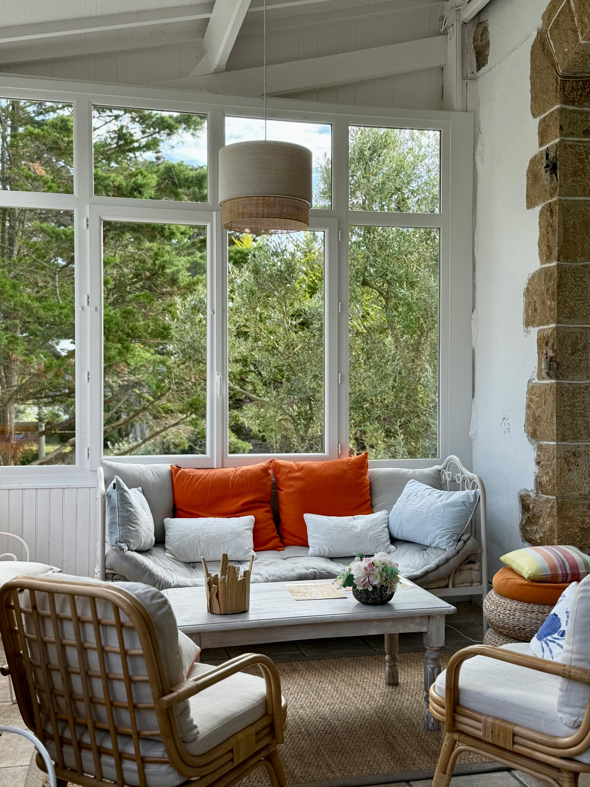 Véranda lumineuse avec mobilier en osier blanc, coussins orange et gris et grandes fenêtres donnant sur la verdure.