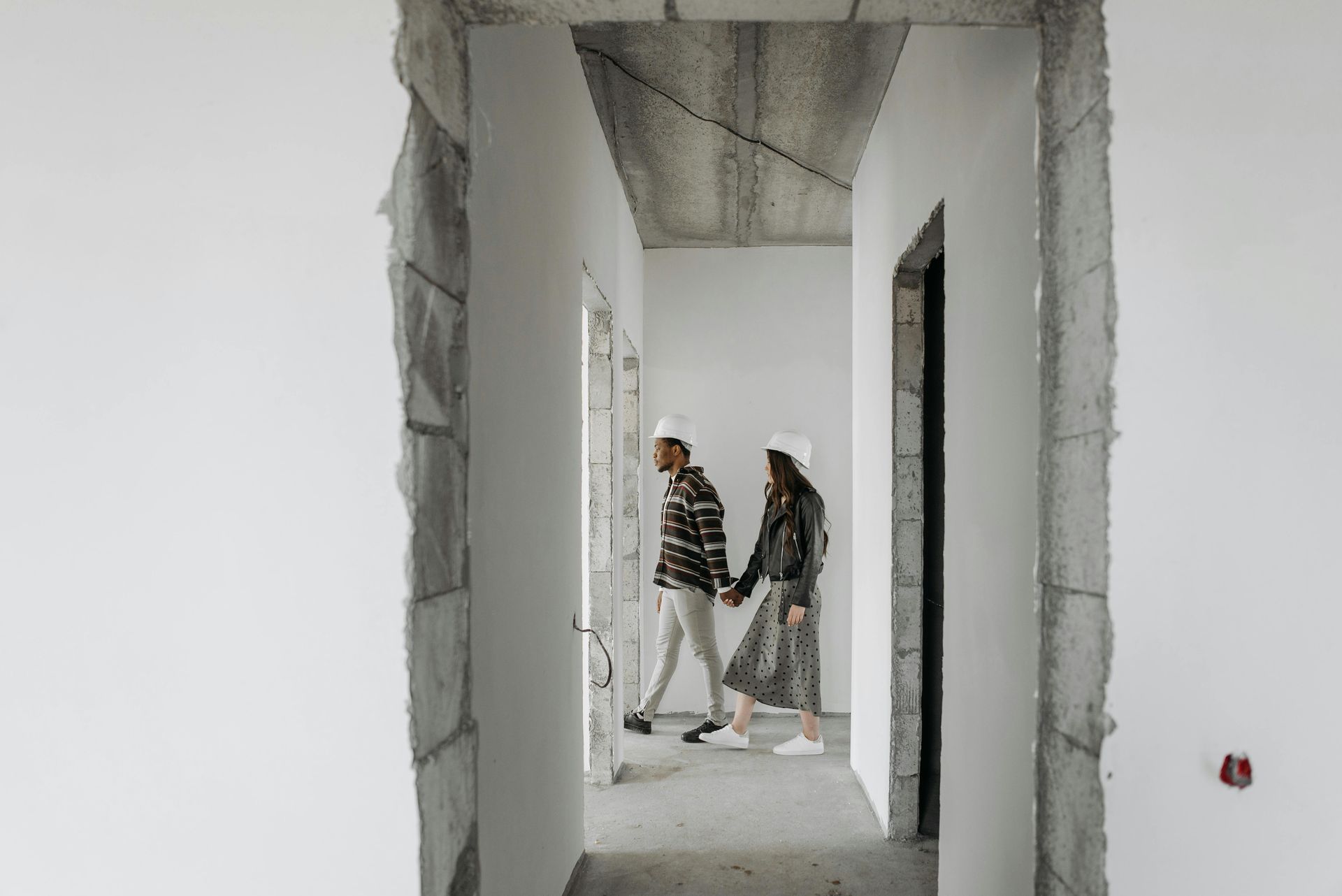 Deux personnes dans un couloir inachevé, traversant une porte en béton brut dans un intérieur blanc.