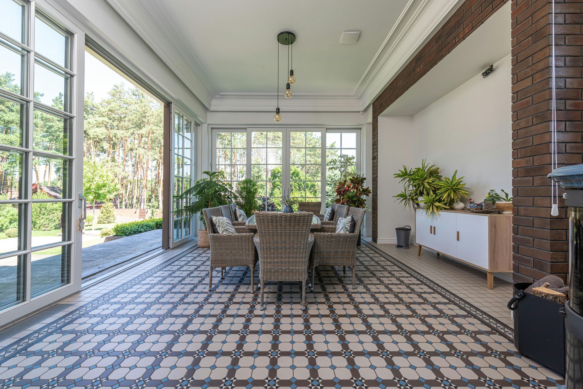 Terrasse couverte avec coin repas, sol carrelé à motifs, chaises en osier et grandes fenêtres donnant sur la verdure.