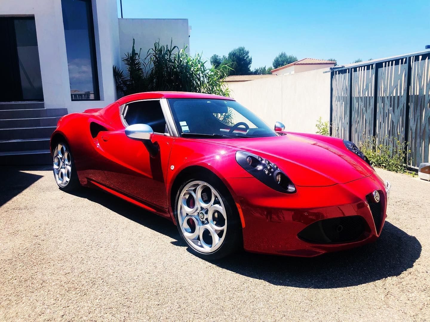 Une Alfa Romeo 4C rouge, voiture de sport, est garée sur une allée de gravier devant une maison moderne.