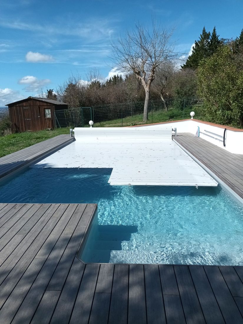 Piscine avec couverture blanche fermée, entourée d'une terrasse en bois, sur un fond vallonné et ensoleillé.
