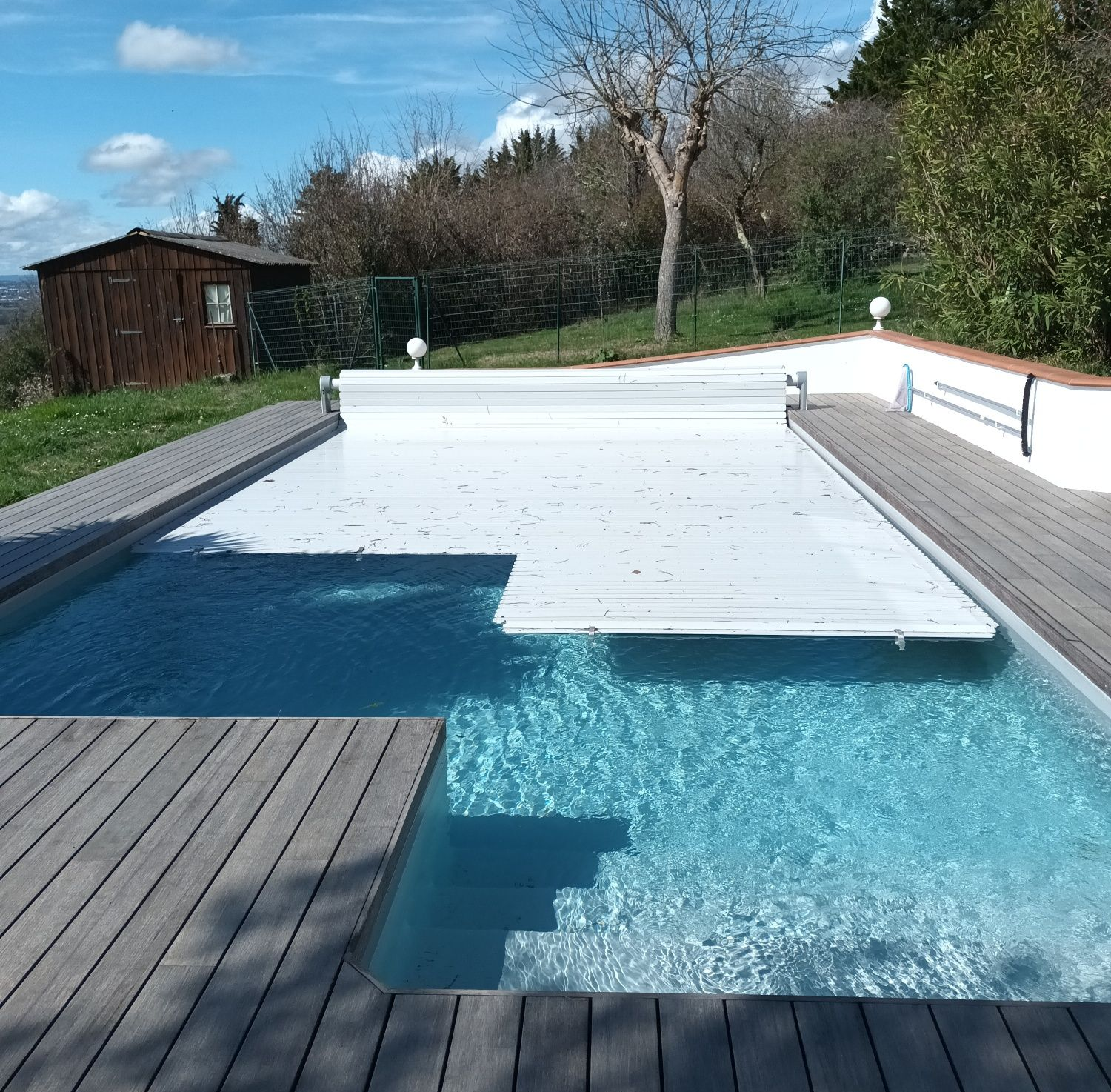 Une piscine avec couverture automatique blanche partiellement couverte, entourée d'une terrasse en bois et d'un petit abri.